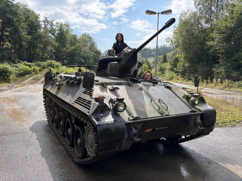 Zwei Gäste auf dem Kettenpanzer, einer winkt aus dem Fahrerplatz
